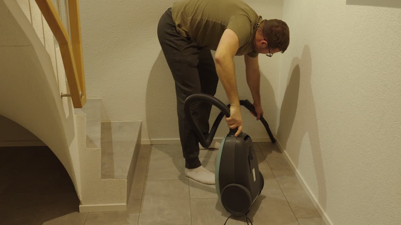 A man struggles to clean floor corners using an old vacuum cleaner. Traditional cleaning concept, hard work, and household routine