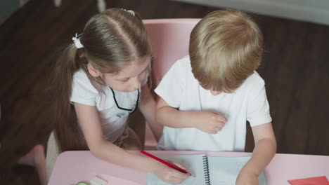 Caring-sister-draws-picture-for-brother-at-desk-at-home