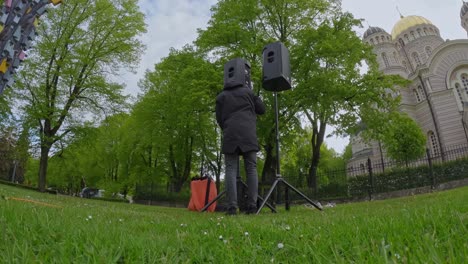 technician installs large speakers on tripods in a green park during an outdoor event setup, captured in wide-angle time-lapse, with historical stone building in background and spring foliage