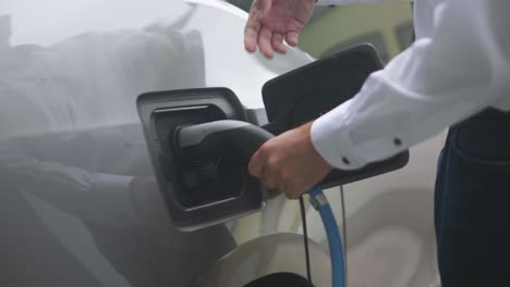 primer plano de un cargador de coche eléctrico moderno conectado a un coche eléctrico plateado por una persona que lleva una camisa blanca