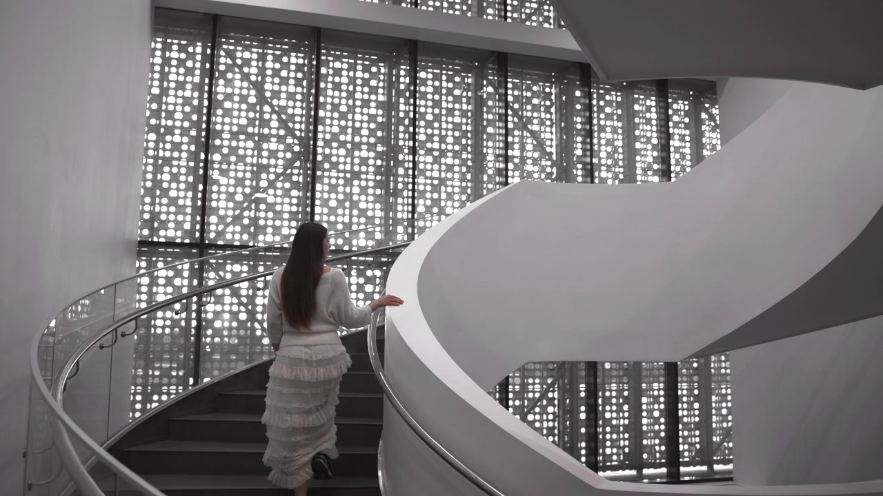 Rear view of woman ascending curved white spiral staircase, one hand touching stainless steel handrail inside modern minimalist interior with perforated backlit window wall, soft light, and clean