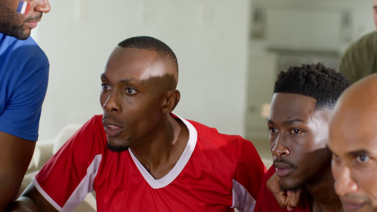 Vertical video: Diverse male fans leaning on sofa in red kit holding soccer ball, reacting to match
