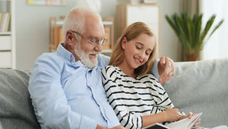 Portrait-Shot-Of-The-Grandfather-In-Glasses-And-Cute-Teen-Granddaughter-Reading-A-Book-Together-While-Sitting-On-The-Couch