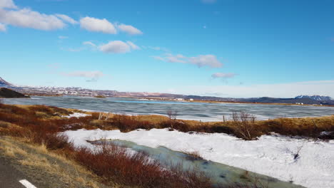 vista desde el coche en el lago myvatn con vista a las montañas, el lago medio congelado y reluciente y la hierba y los arbustos rojos dorados