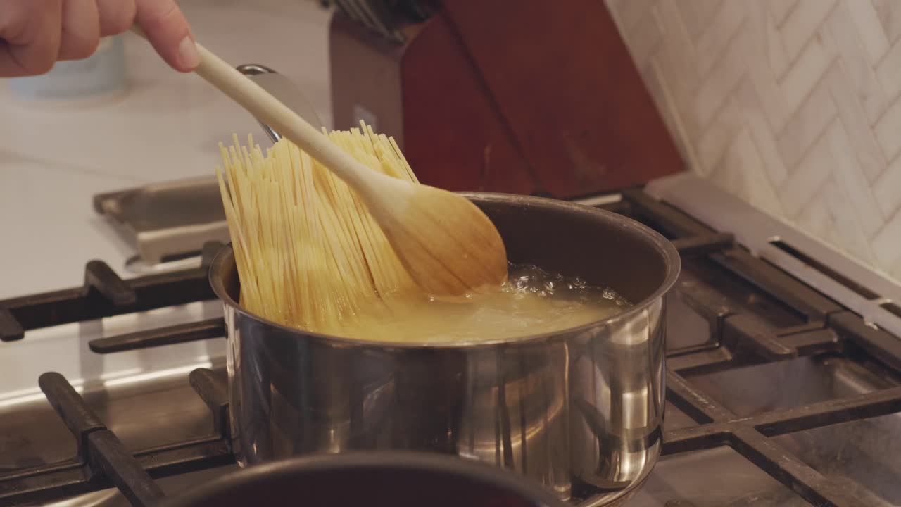 Un primo piano al rallentatore di un cucchiaio di legno che mescola la pasta mentre si ammorbidisce in una pentola d'acqua bollente. Ripresa in una moderna cucina domestica