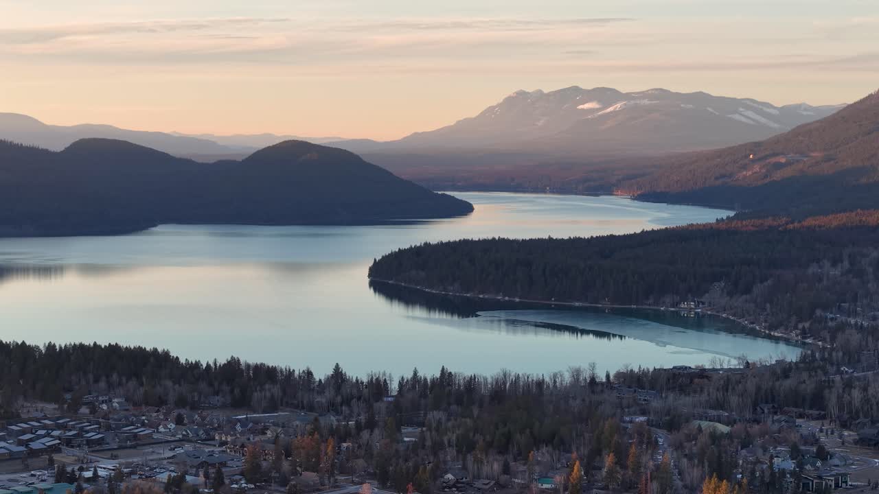 Riprese aeree con drone mostrano un lago sinuoso delimitato da una foresta di conifere e distanti montagne innevate vicino a Whitefish, Montana, all'alba. Nessun animale visibile