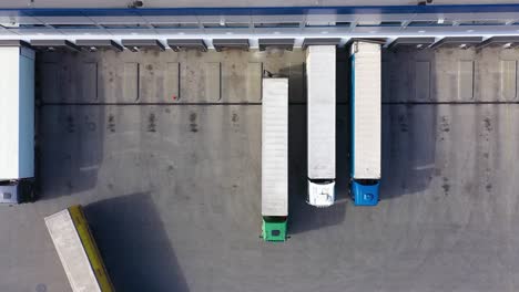 Aerial-Shot-of-Truck-with-Attached-Semi-Trailer-Leaving-Industrial-Warehouse-Storage-Building-Loading-Area-where-Many-Trucks-Are-Loading-Unloading-Merchandise