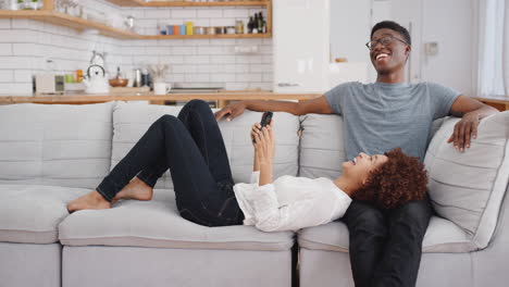 pareja joven relajándose en el sofá en casa mirando el teléfono móvil y riendo juntos