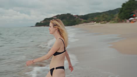 happy young blonde woman in black swimsuit walking along sandy beach into sea in thailand