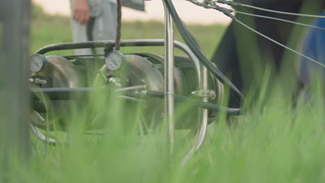 close up of hot air balloon burner system rotating beside tall green grass moving in wind as blurred figure stands in background during outdoor setup on launch site with visible cables and pipes