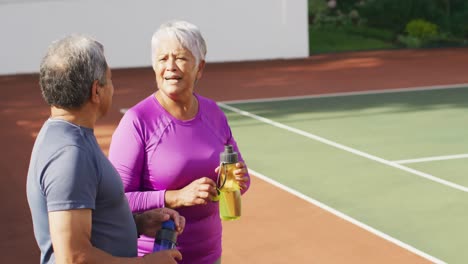 video of happy biracial senior couple drinking water and talking after training on tennis court