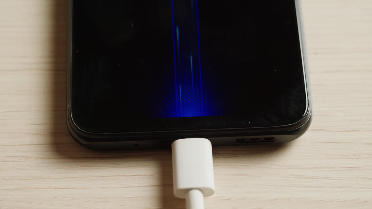 A smartphone connected to a USB cable is charging on a table, surrounded by tech accessories and devices in a photo