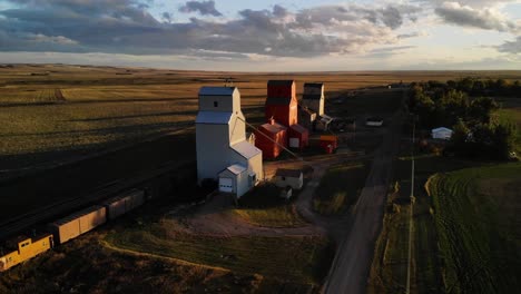 Tiro-Giratorio-Aéreo-De-Tres-Elevadores-De-Granos-Rodeados-De-Campos-Agrícolas-En-Un-Día-Nublado-En-Alberta,-Canadá-Al-Atardecer