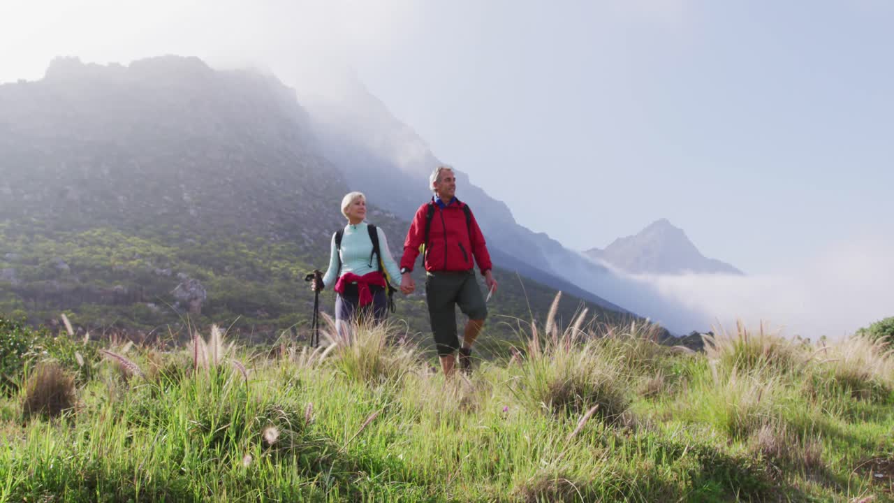 Coppia di escursionisti anziani con zaini e bastoncini da trekking che si tengono per mano e camminano mentre fanno trekking.