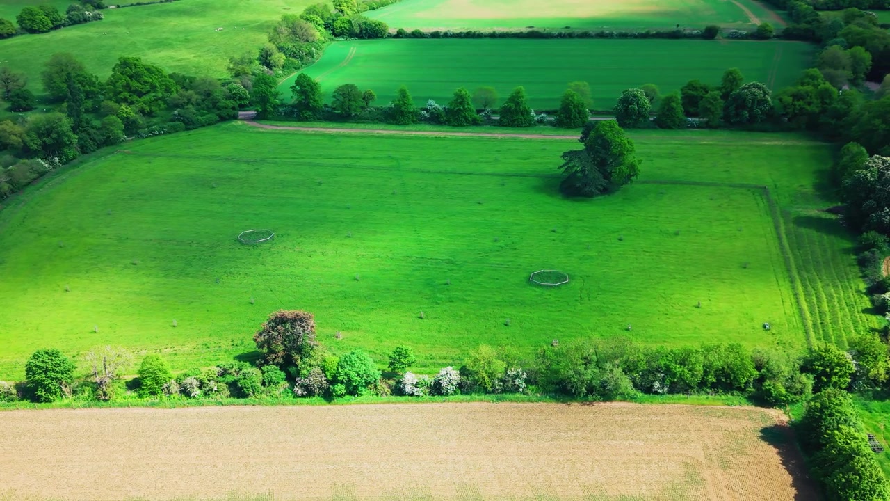 Riprese aeree con drone su campi di pascolo verdi, siepi, alberi e terreni agricoli circostanti nella campagna inglese. L'inquadratura mostra confini di campi rurali, margini di boschi, terreno agricolo e un paesaggio rurale