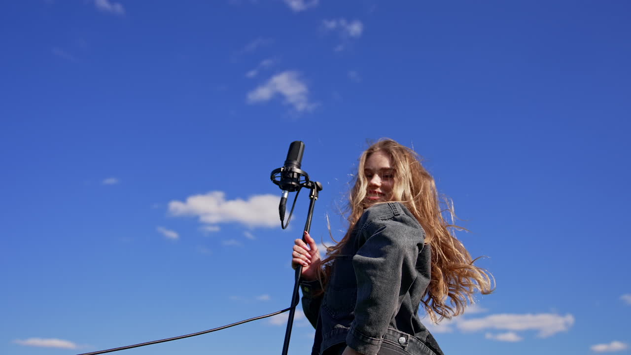 Pop singer woman on sky background. Beautiful girl in jeans clothing dancing and singing into the microphone on field in sunny day.