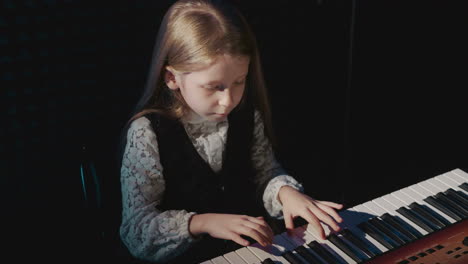 child plays simple melody on electric piano keyboard. blonde little girl in formal clothes performs musical melody in audio studio. acoustic concert