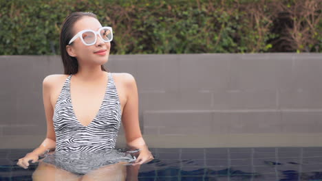 Medium-close-of-a-young-woman-in-a-zebra-pattern-one-piece-swimsuit,-lounging-in-the-shallow-end-of-a-resort-swimming-pool