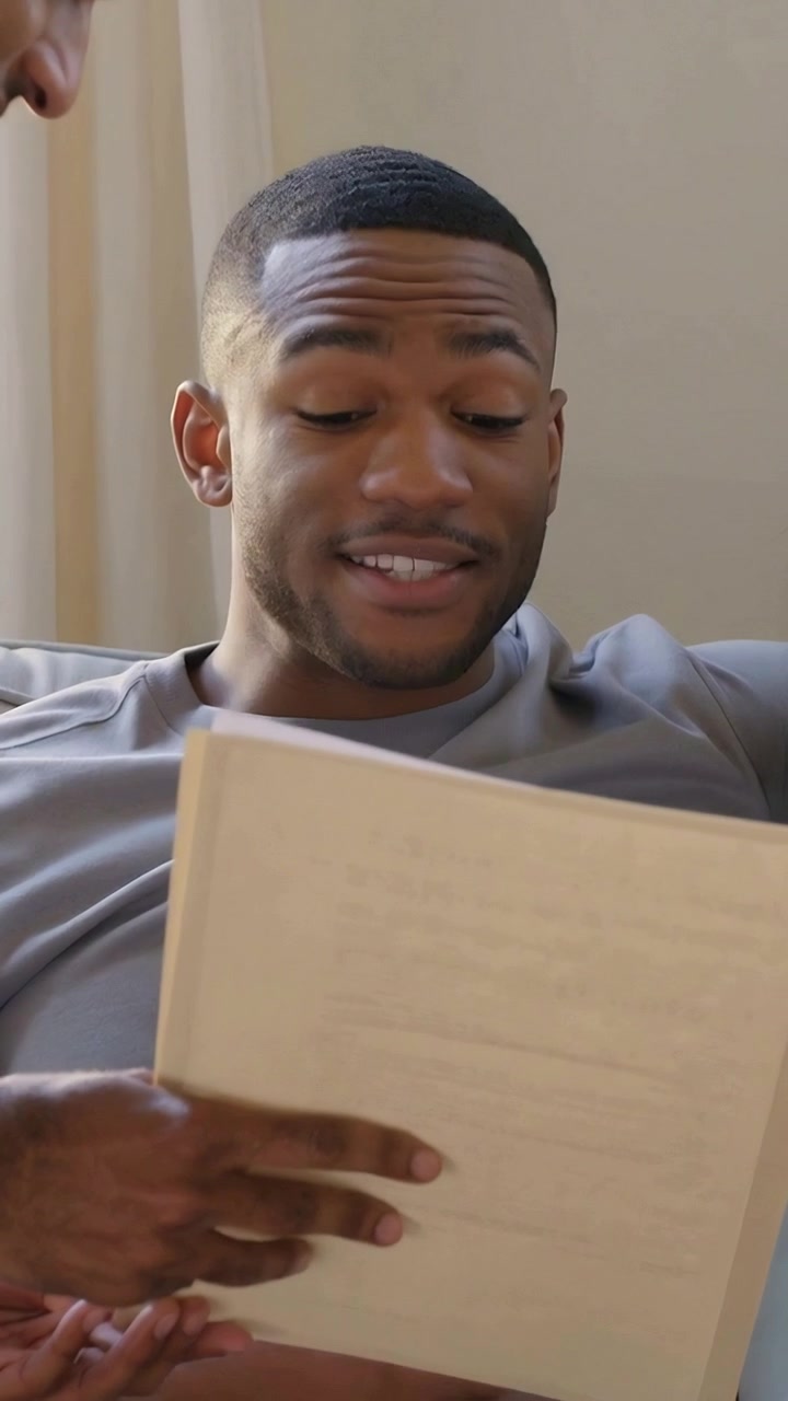 Vertical video: Leaning man pointing to notes at home, seated man in gray shirt reading, explaining
