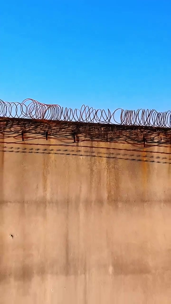 Video verticale: Telecamera in panoramica che monitora un muro perimetrale, rivelando filo spinato a spirale e staffe.