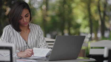 Business-Woman-Brunette-Hispanic-Ethnicity-Works-remotely-while-sitting-in-a-summer-cafe-on-a-sunny-day-with-a-laptop-and-writes-down-with-a-pen-and-notebook