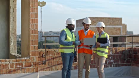 caucasian woman and mixed-races men standing together in hardhats at the building site with laptop computer, workday of builders, constructors and architects.