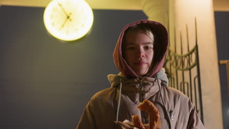 elegant girl standing with pastry in hand in a busy urban setting, cars moving in background under streetlights, cozy yet modern atmosphere, with a glowing night clock in the background