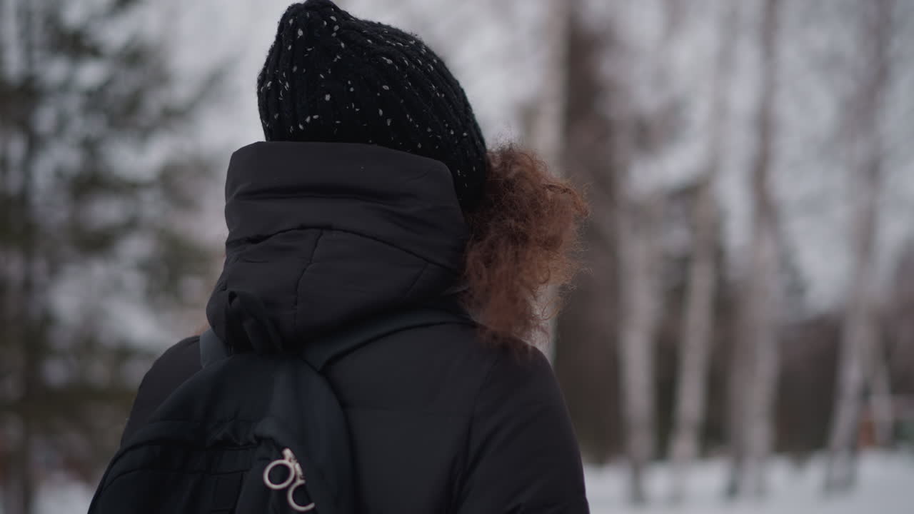 Mujer de pie en un parque nevado en invierno vista desde atrás con abrigo negro, mochila, gorro, pelo rizado, soledad, estación fría al aire libre, ambiente gélido, entorno congelado, paisaje tranquilo, cielo cubierto, calma