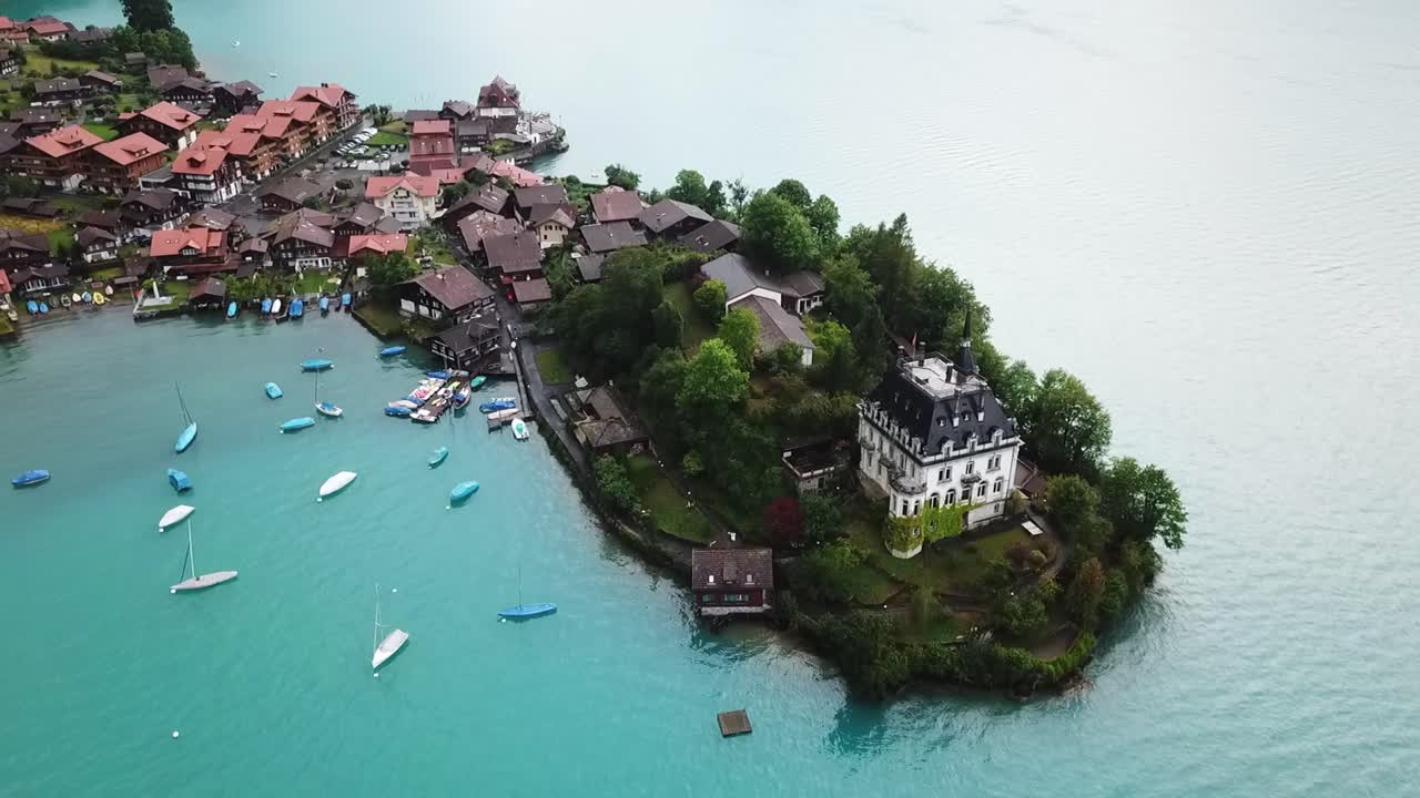 Aerial view of Iseltwald, a picturesque village on Lake Brienz, Switzerland, with charming houses, lush greenery, and boats docked along the turquoise water as the drone descends