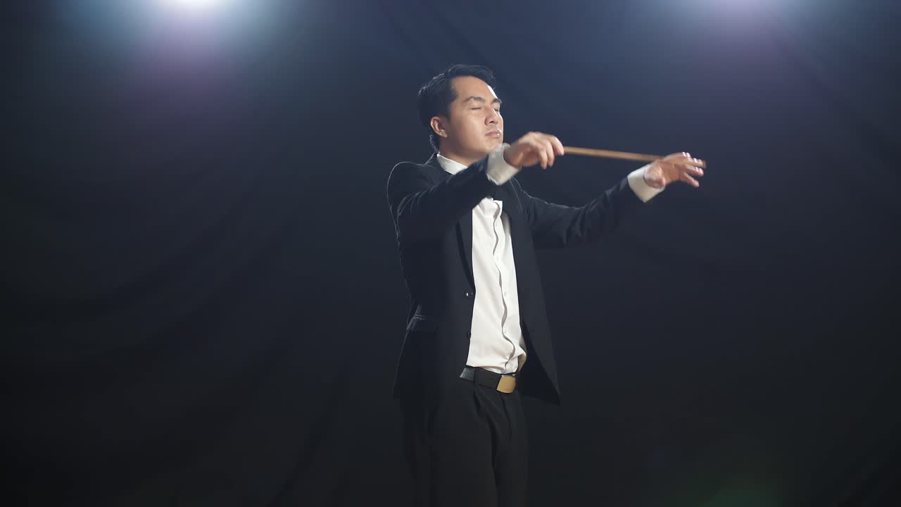 Side View Of Asian Conductor Man Holding A Baton Closing His Eyes And Showing Gesture In The Black Studio