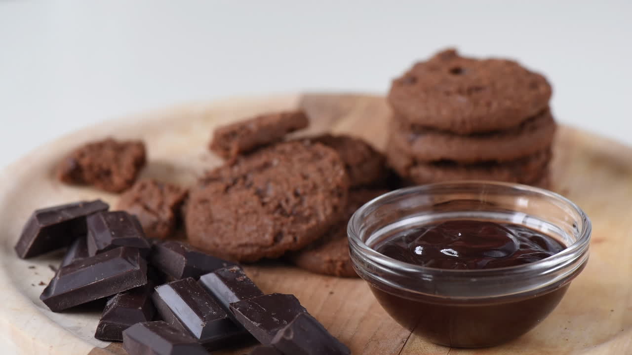 Close up of a woman dipping a cookie in a chocolate paste near pieces of dark chocolate on a wooden tray