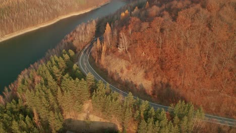 an aerial ascending drone view of cars driving on a winding road alongside a lake in late autumn