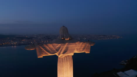 Close-up-dolly-out-aerial-night-shot-of-Santuario-de-Cristo-Rei-white-monument-on-the-hill-with-city-center-of-Lisbon-with-lights-in-the-background