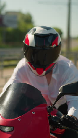 a female motorcyclist wearing a helmet rides a red power bike fast near a walk path with an iron rail, with a blur lush green tree in view by the side