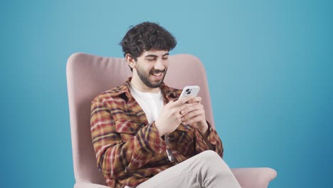Young-man-sitting-on-sofa-texting-on-phone,-smiling,-texting-on-social-media.