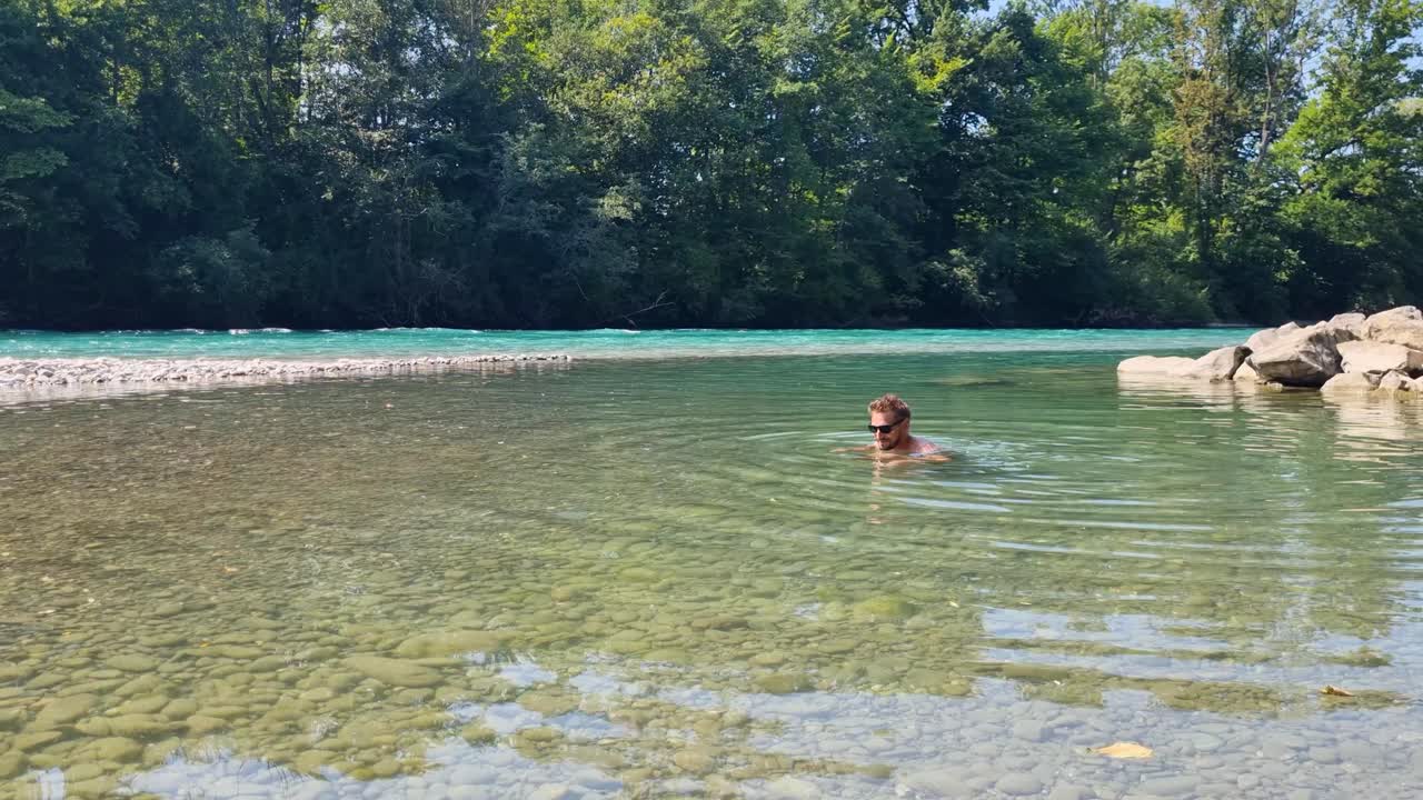 Uomo attraente nuota e si rilassa nelle acque turchesi del fiume Aare, godendosi una soleggiata giornata estiva in mezzo a una natura verde lussureggiante e un paesaggio rinfrescante.