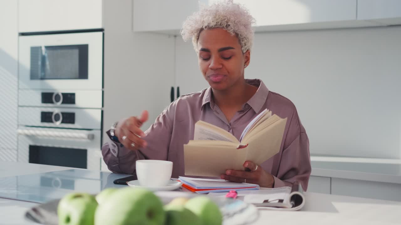 joven mujer afroamericana bostezando leyendo un libro en el desayuno se sienta en la cocina