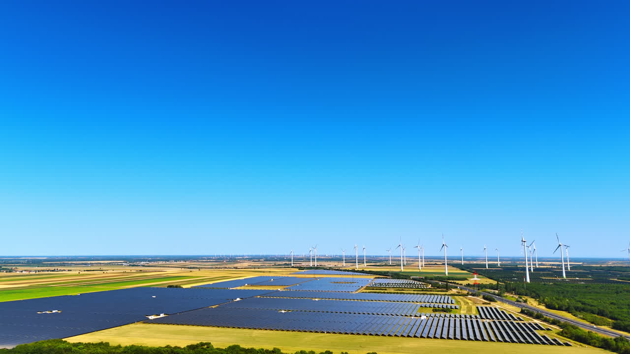 Renewable energy fields under clear sky. Expansive solar panels and wind turbines stretch across farmland under a bright blue sky on a sunny day
