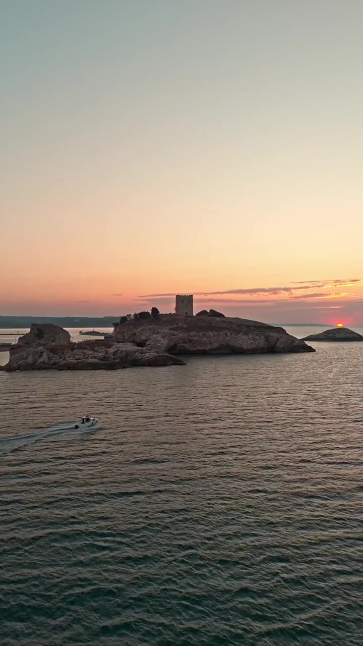 Dronebillede der viser en båd passere If Slot under solnedgang nær Marseille, Frankrig. Gyldent lys oplyser den klippefyldte ø og det rolige Middelhav, hvilket skaber et naturskønt kystlandskab.