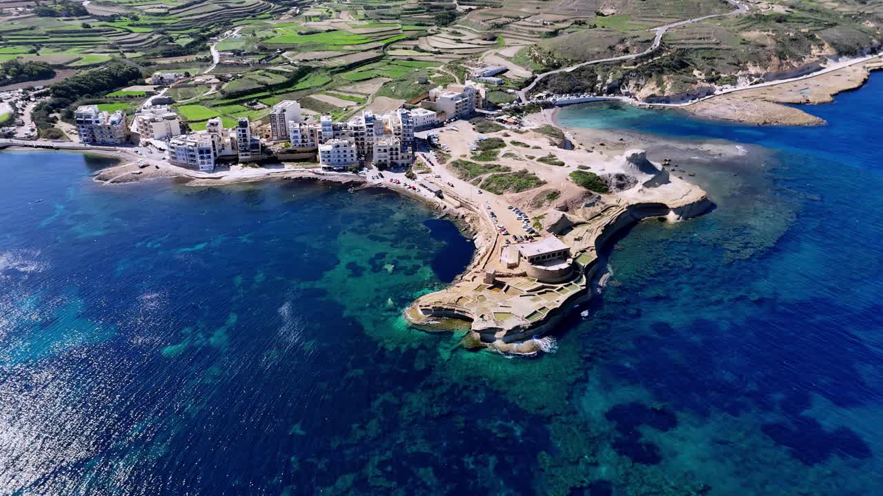 Vista aerea dall'alto di Marsalforn Bay, Gozo, Malta, sotto il vibrante sole estivo, con acque turchesi del Mediterraneo, costa rocciosa calcarea, campi terrazzati verdeggianti e moderna architettura costiera