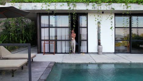 Shirtless-Man-Walks-Out-From-Tropical-Luxury-Villa-to-Private-Infinity-Swimming-Pool-While-Drinking-Morning-Cup-of-Coffee,-Aerial-Push-Back