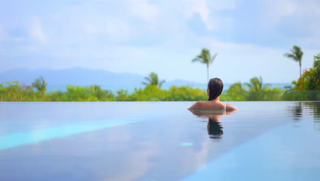 vista trasera de una mujer nadando dentro de la piscina de un hotel tropical con palmeras y panorama de montañas en el fondo en bali