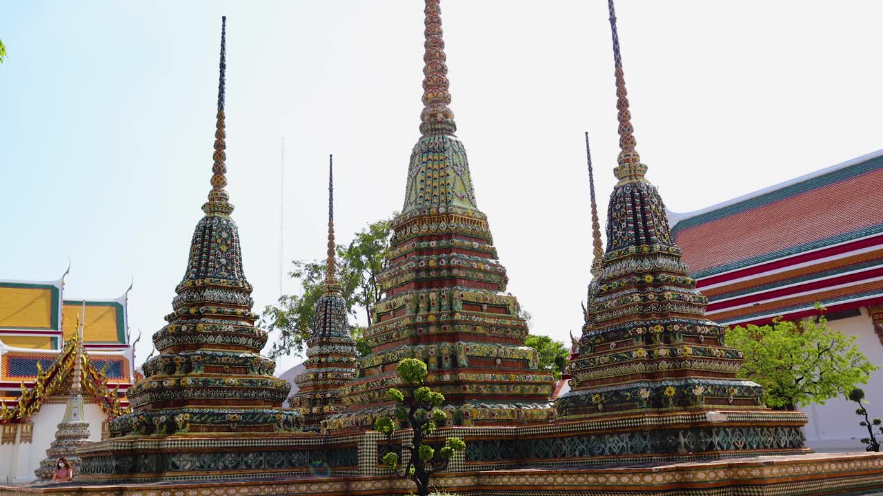 A tranquil scene of ornate stupas at Wat Pho, Bangkok, under clear blue skies. The camera pans upward, highlighting architectural details