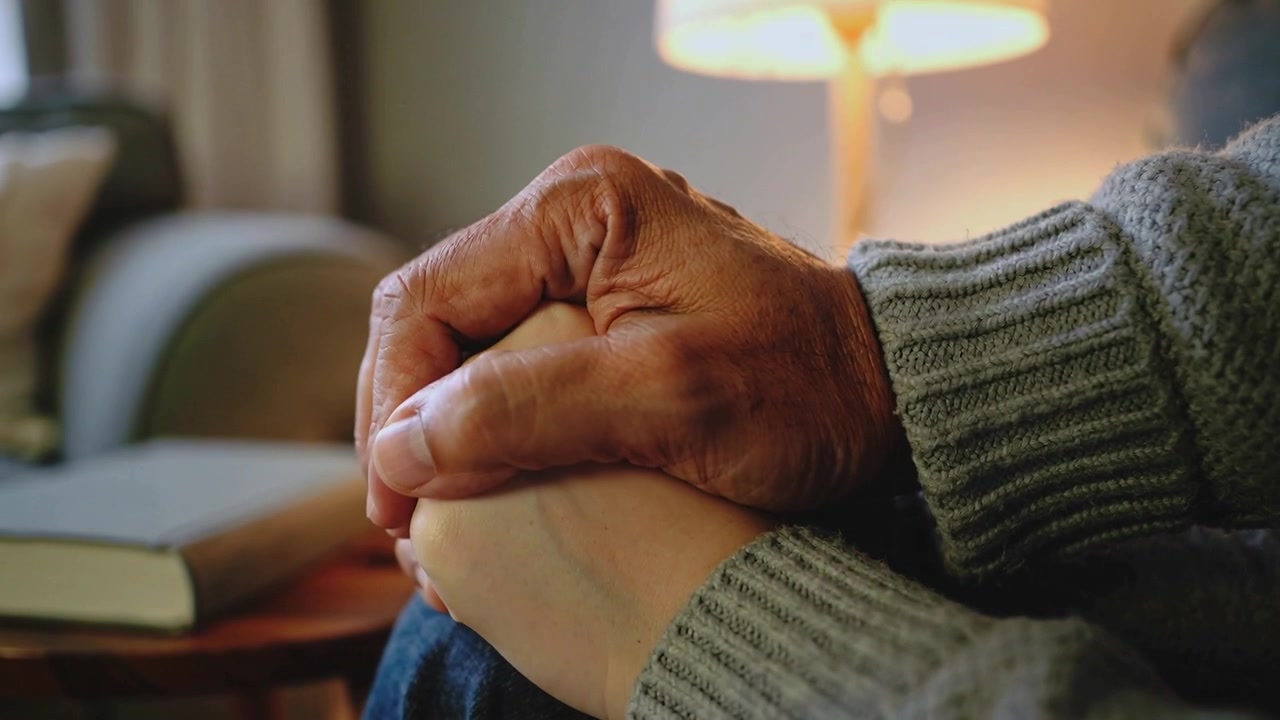Camera framing wrinkled hand squeezing lighter hand in living room, knit sleeves, lamp comforting
