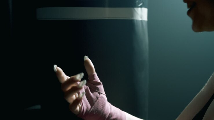 Primo piano delle mani di una donna bionda con unghie lunghe che srotola bende rosa per le mani dopo l'allenamento. Si trova vicino a un sacco da boxe. L'illuminazione cinematografica evidenzia la texture, il movimento e l'atmosfera della palestra
