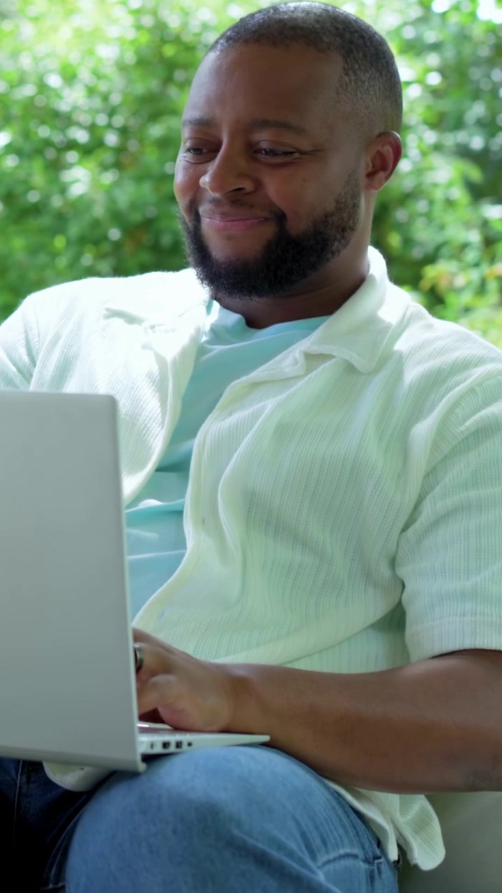Vertical video: Sitting man wearing overshirt and blue tee typing on laptop on patio, working