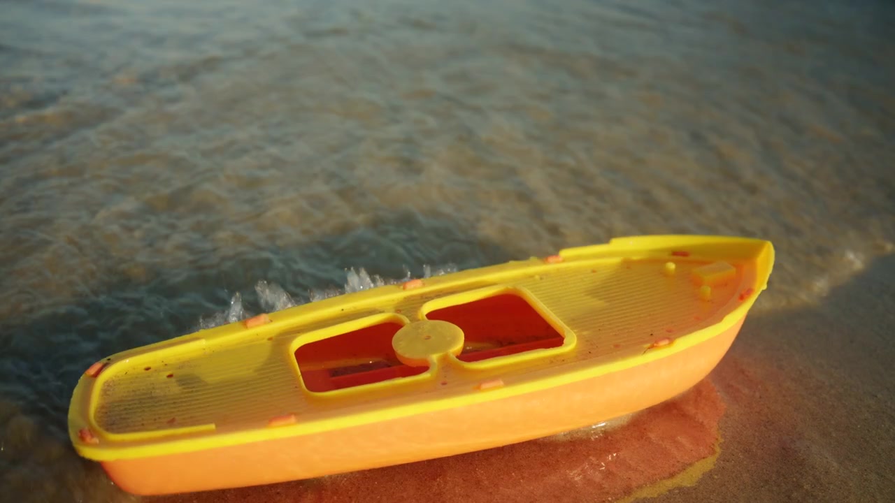 Barca giocattolo di plastica che riposa sulla riva sabbiosa mentre dolci onde la lambiscono, simboleggiando il gioco infantile e l'avventura estiva.