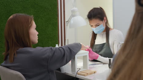 nail technician wearing protective gloves carefully filing customer nail at salon desk while client gazes upward, with visible salon tools, green wall panel, and partial view of another blonde hair