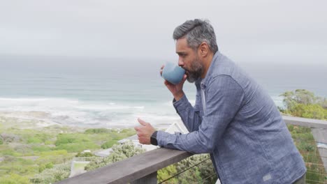 Biracial-man-drinking-mug-of-coffee-and-thinking-on-balcony-alone