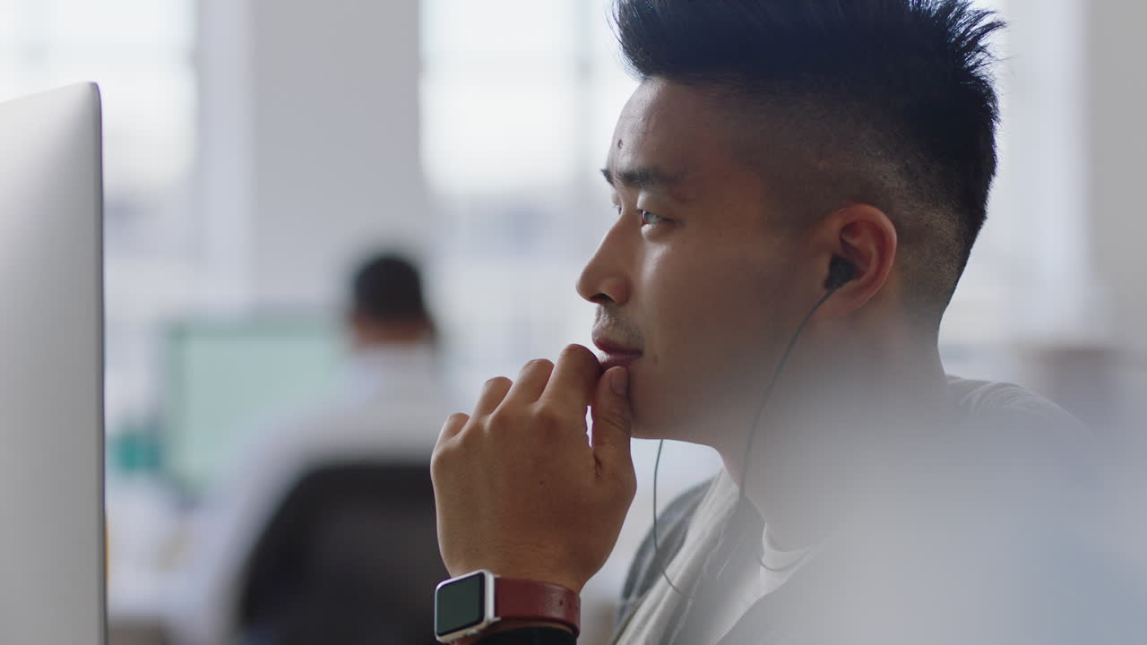 young asian businessman student using computer brainstorming problem solving idea looking pensive thinking of solution in office workplace wearing earphones